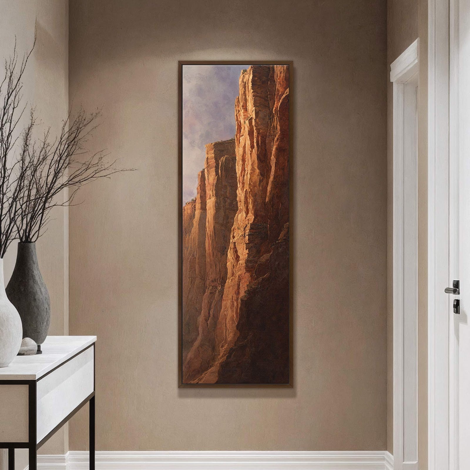 This vertical canvas print showcases a realistic painting of sunlit canyon cliffs with a warm, earthy palette, framed in dark wood, and displayed on a taupe wall above a console.