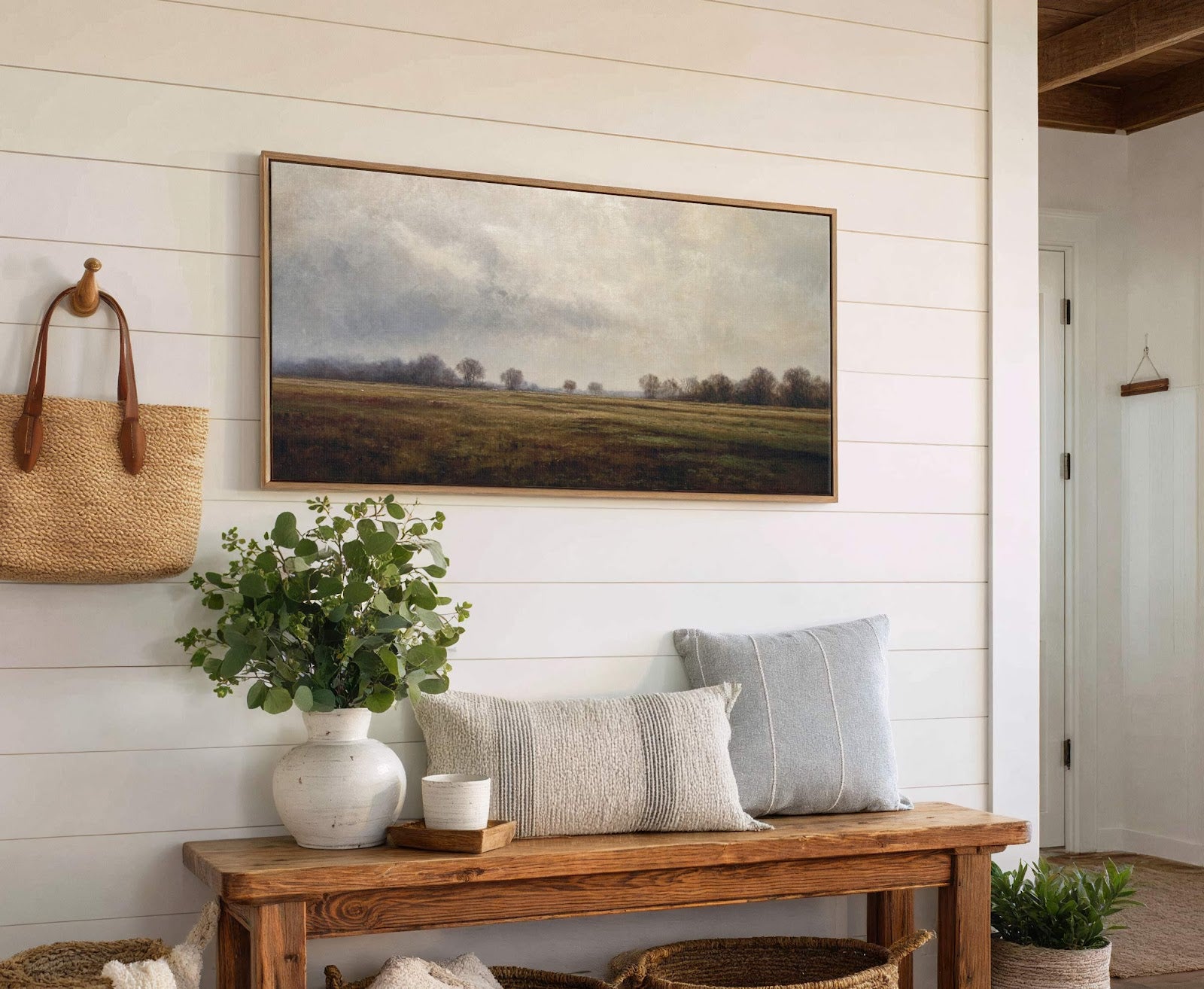 This horizontal canvas wall art depicts a muted landscape with trees and fields under a cloudy sky, framed in natural wood, and hangs above a wooden bench with pillows and.