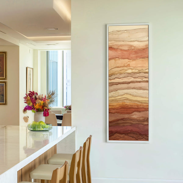 A vertical, abstract artwork print featuring horizontal bands of earthy browns and oranges with thin, dark lines, framed in white, hangs on a light cream wall above bar stools in.