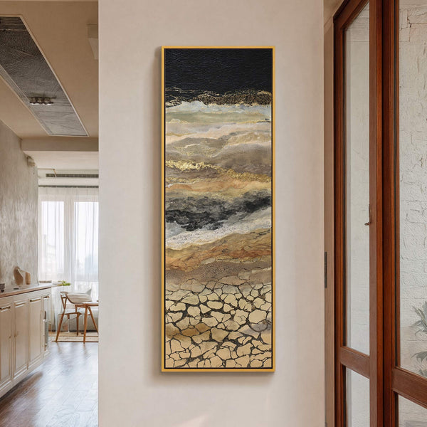 This vertical abstract canvas wall art features a textured palette of black, gold, beige, and gray, evoking a cracked earth or layered landscape. It hangs on a light-colored wall near.