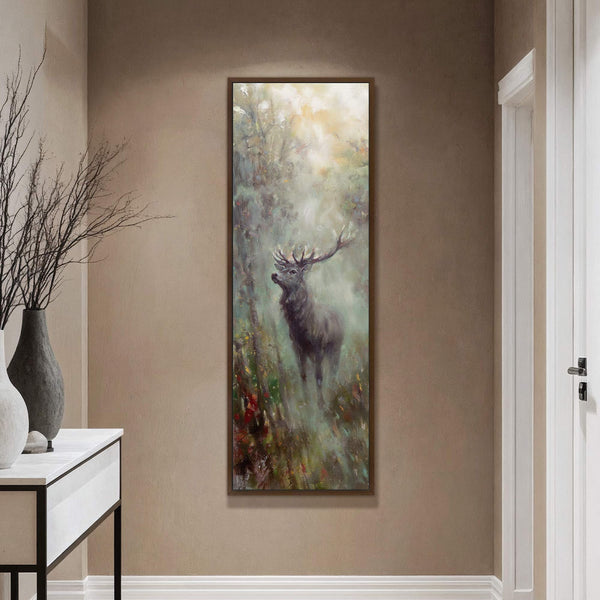 This vertical canvas wall art depicts a stag in a forest with a misty, impressionistic style and a warm, earthy palette, displayed on a tan wall next to a console.