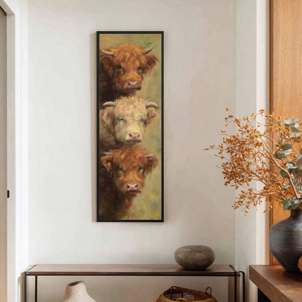 This vertical canvas wall art features a painterly style with a warm, earthy palette depicting three Highland cows stacked vertically, framed in black and hanging above a wooden console table.