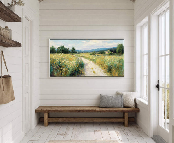 This horizontal canvas wall art features a painterly landscape with a dirt path through a field of wildflowers and distant mountains, framed in white and displayed in a room with.