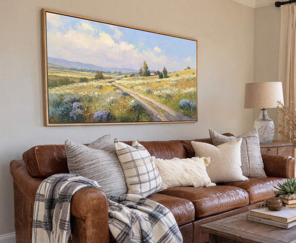 A horizontal landscape painting print with impressionistic brushstrokes depicts a sunny, rural path winding through fields of wildflowers, with mountains in the distance. The artwork is framed in natural wood.