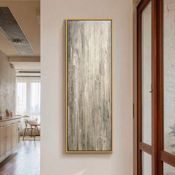 This vertical abstract art print features a palette of gray and off-white with a textured, streaky style. It is framed in gold and hangs on a light-colored wall in a.
