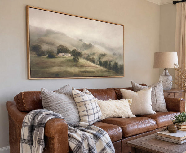 This horizontal canvas wall art features a muted palette and impressionistic style, depicting a misty landscape with a small house. It is displayed above a brown leather sofa with decorative.