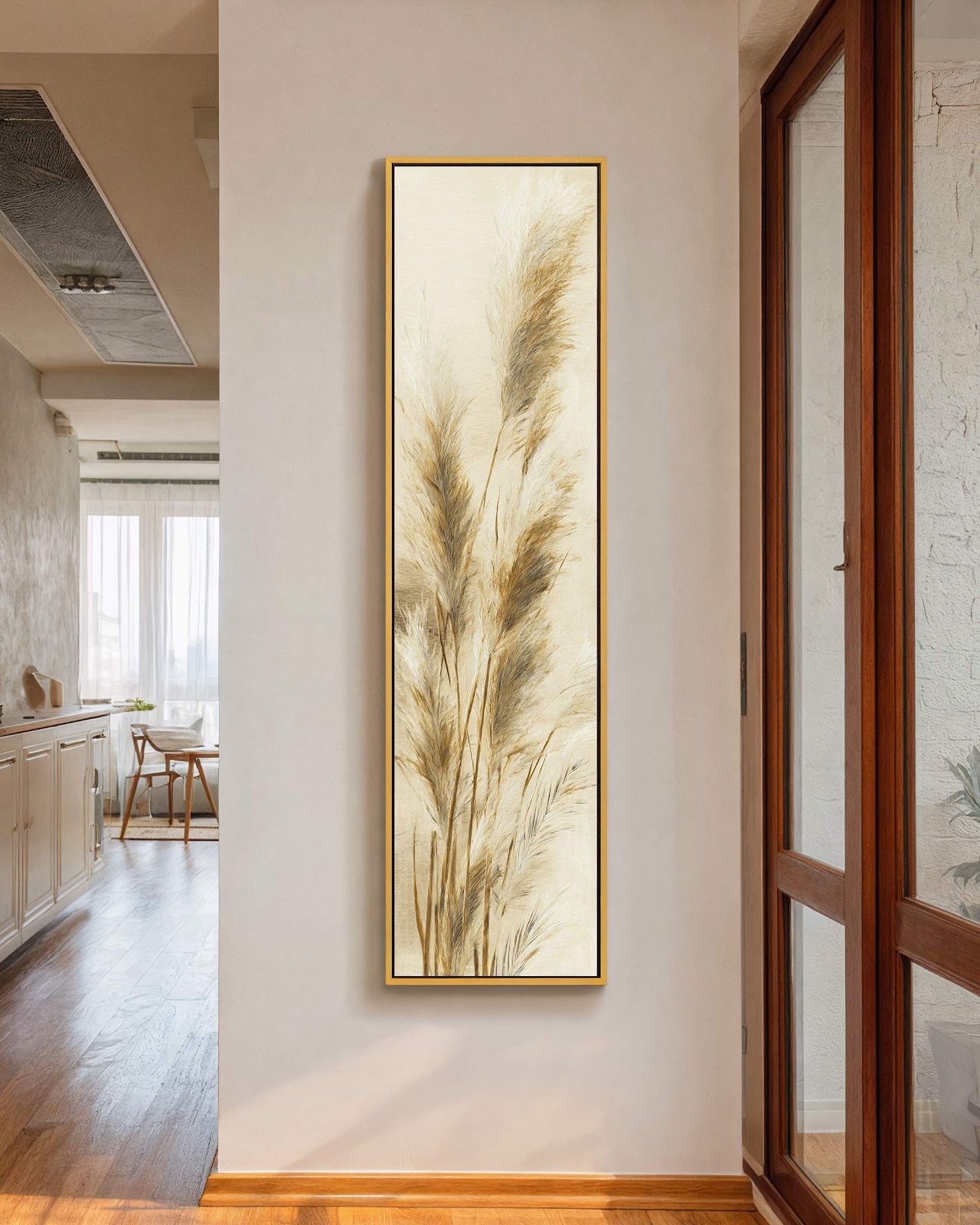 This vertical canvas print displays textured pampas grass in a neutral palette with a gold floater frame, hanging on a white hallway wall near a large window.