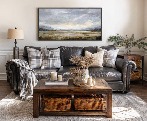 A horizontal canvas wall art print with a muted landscape painting in gray and gold tones, framed in black, hangs above a dark leather sofa on a white wall.