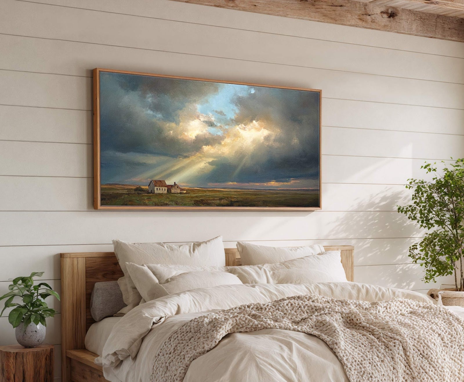 This horizontal canvas wall art features a dramatic landscape painting with stormy clouds and sunbeams illuminating two houses in a field, framed in natural wood and displayed above a bed.