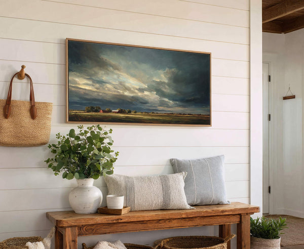 This horizontal canvas wall art features a dramatic landscape with dark, stormy clouds over fields and red barns, set against a white shiplap wall with a wooden bench and pillows.