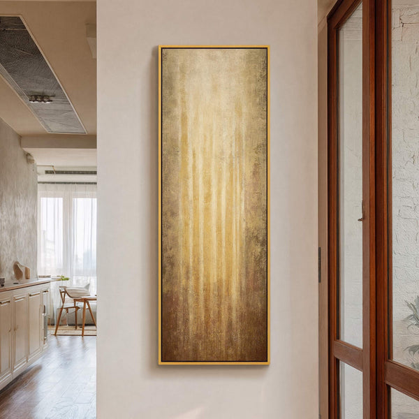 This vertical abstract art print features a warm, textured palette of golden browns and yellows, framed with a thin gold border and displayed on a neutral wall within a hallway.