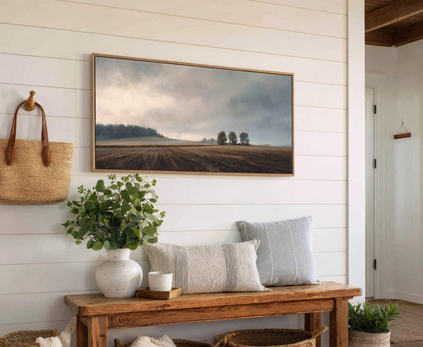 This horizontal canvas wall art depicts a somber, misty landscape with a field and trees under a cloudy sky, framed in natural wood, and displayed above a wooden bench with.