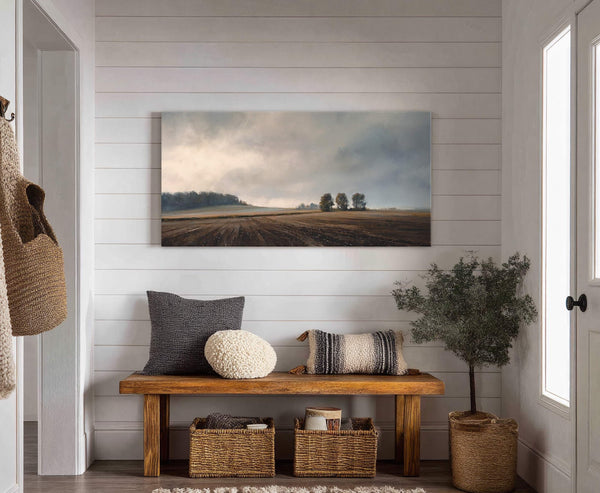 A horizontal canvas print displays a muted landscape with a cloudy sky over plowed fields and distant trees, set on a white shiplap wall above a wooden bench with pillows.