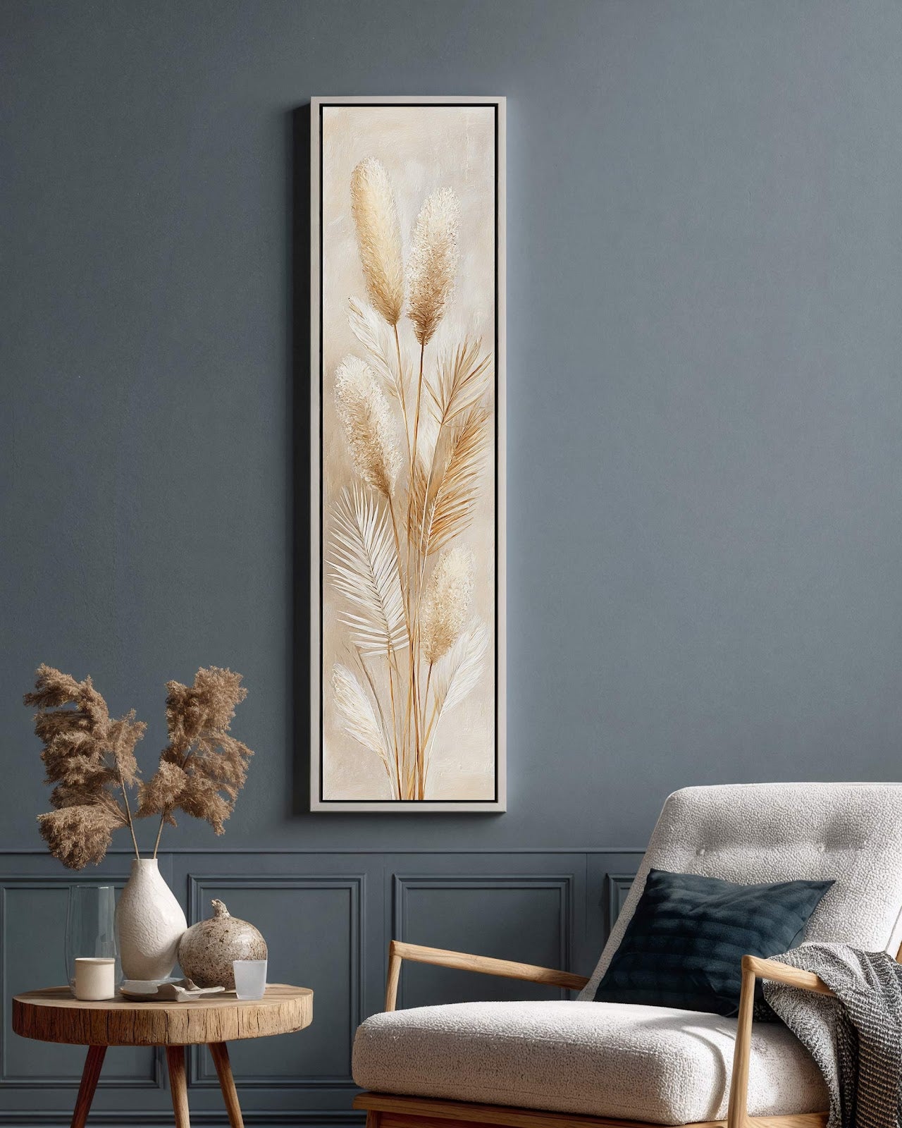 This vertical canvas print depicts textured pampas grass in beige tones, set in a white floater frame on a blue paneled living room wall near a white armchair.