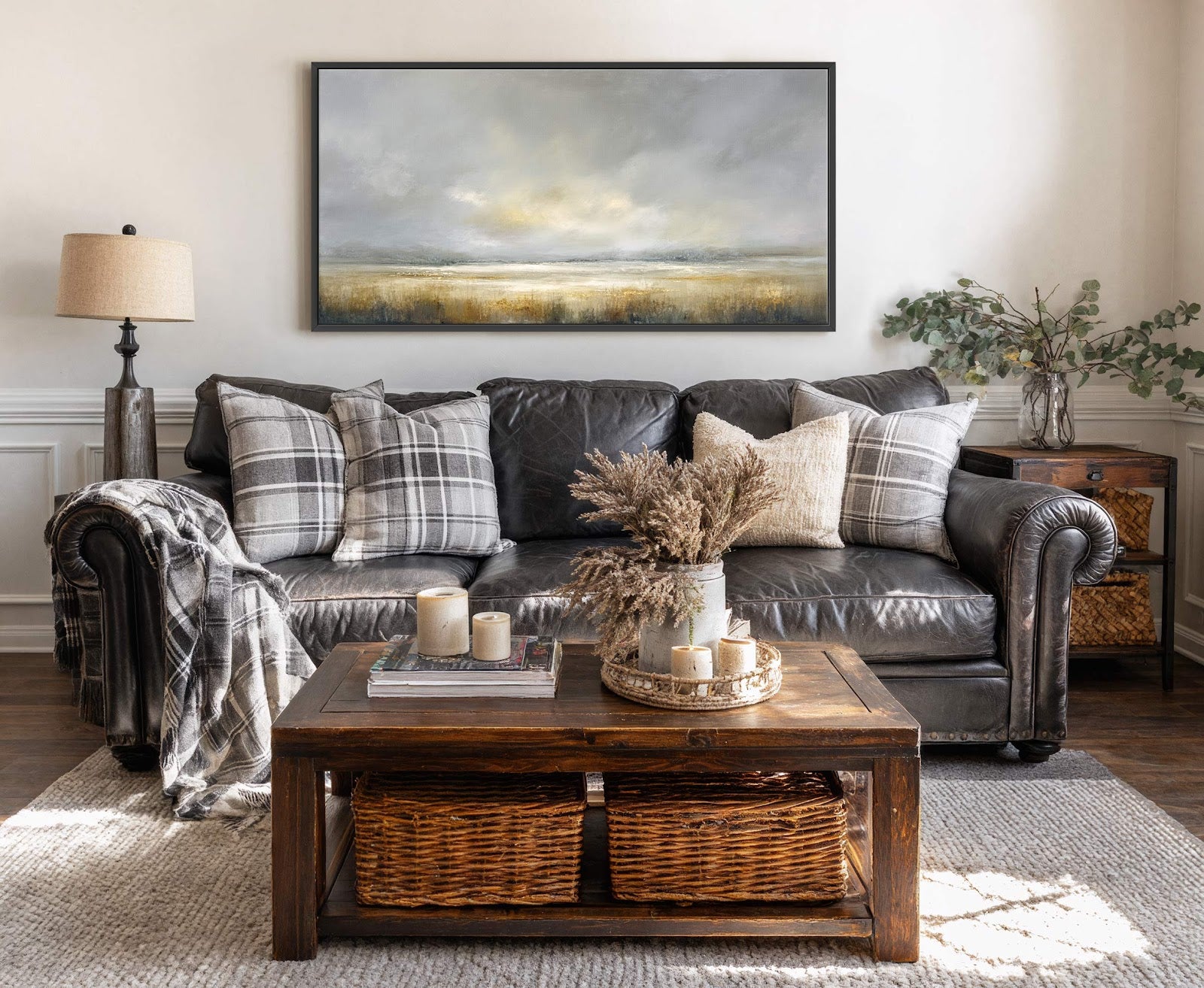 This horizontal canvas wall art features an atmospheric landscape painting with a muted palette of grays, blues, and ochres, displayed above a dark leather sofa with plaid pillows in a.