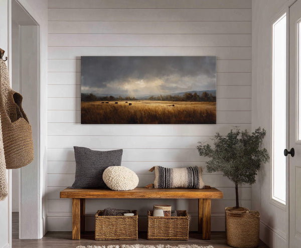 This horizontal canvas wall art depicts a moody landscape with dramatic clouds over a golden field of hay bales and trees, hanging on a white paneled wall above a wooden.