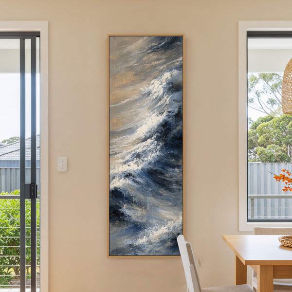 This vertical, abstract expressionist painting print depicts stormy ocean waves in a cool blue and neutral palette, framed in light wood and displayed on a cream-colored wall near a window.