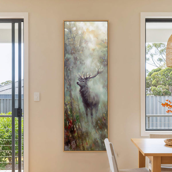 This vertical canvas print displays an impressionistic painting of a stag in a misty forest, rendered in a muted, earthy palette, hanging on a beige wall next to a window.