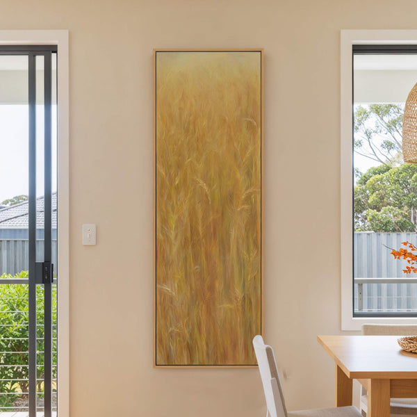 A vertical canvas print displays a golden wheat field in an impressionistic style with warm tones, framed in natural wood, hanging on a pale beige wall near a window and.