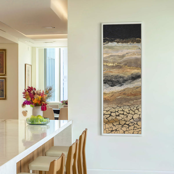 This vertical abstract art print features a palette of black, gold, and earthy tones, with a textured style resembling cracked earth, displayed on a white wall in a room with.