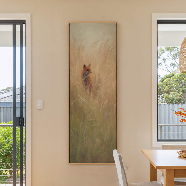 A vertical canvas print depicts a fox in tall grasses with a warm, earthy palette and a loose, painterly style, framed in natural wood, hanging on a beige wall next.