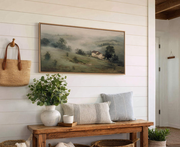 This horizontal canvas wall art showcases a misty, atmospheric landscape painting with a muted green and gray palette, featuring rolling hills and a distant house, displayed above a wooden bench.
