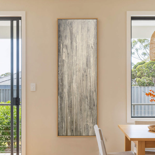 This vertical canvas wall art features an abstract textured design with a palette of gray, white, and beige, displayed on a neutral wall near a window and a dining table.