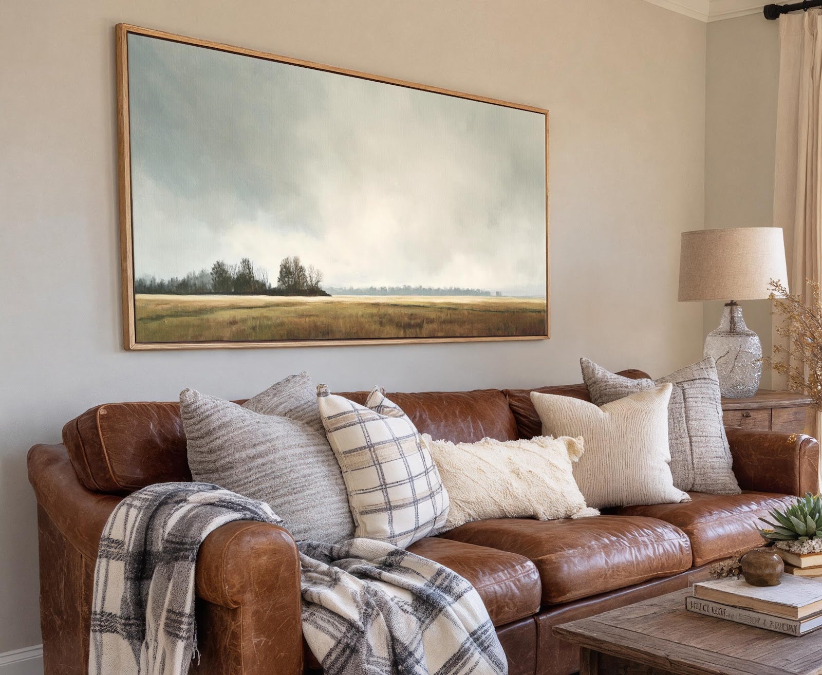 This horizontal canvas wall art displays a muted landscape with a pale blue sky and a hazy field in a painterly style, displayed above a brown leather sofa with pillows.