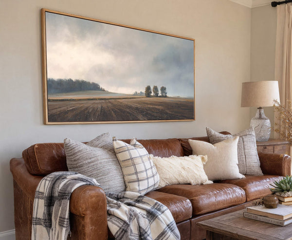 This horizontal canvas print displays a moody landscape painting of a tilled field and trees under a cloudy sky, framed in natural wood, and hangs above a brown leather sofa.
