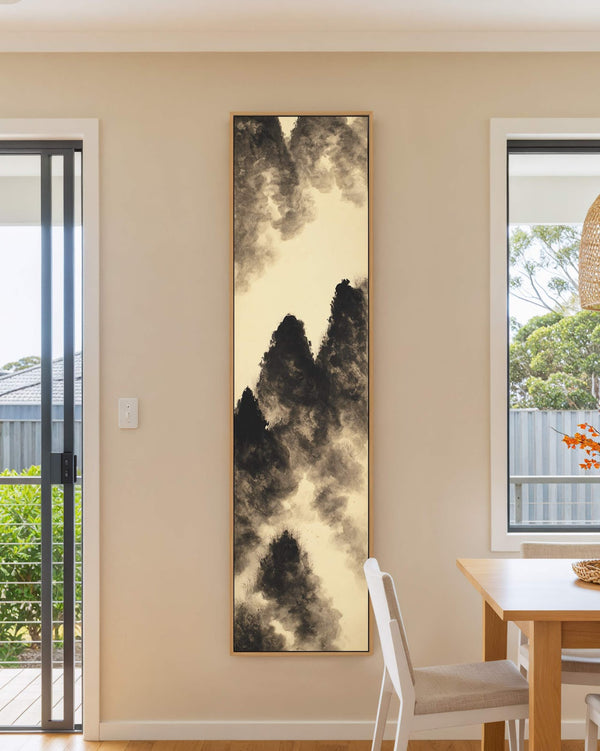 This vertical art print depicts minimalist black ink mountain peaks on a sepia background, hanging in a natural floater frame on a beige dining room wall near a table.