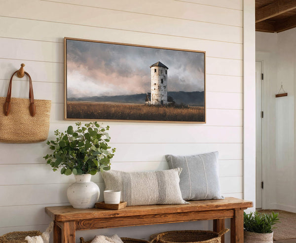 A horizontal canvas print artfully depicts a weathered white tower in a grassy field under a dramatic, cloudy sky with a muted palette, framed in natural wood and displayed on.