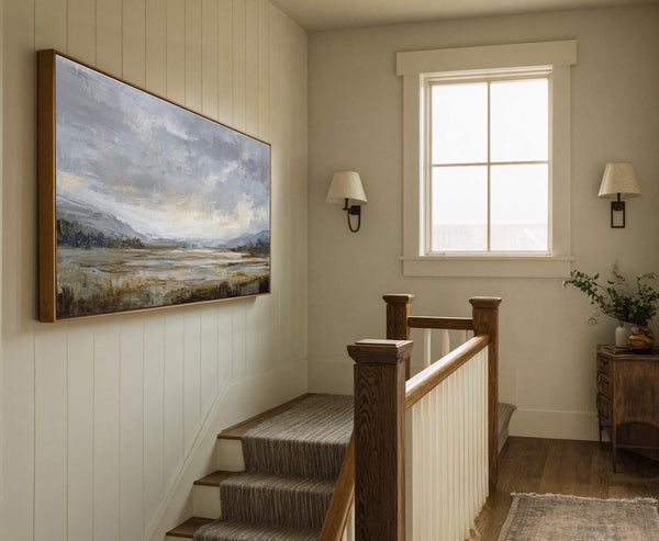 This horizontal canvas wall art features an impressionistic landscape in a muted palette of grays, blues, and browns, framed in natural wood, hanging above a staircase with a tan runner.