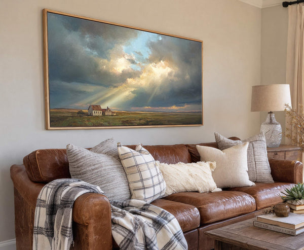 This horizontal canvas print, framed in natural wood, depicts a pastoral landscape under dramatic, light-streaked clouds and a muted palette, hanging above a brown leather sofa in a living room.