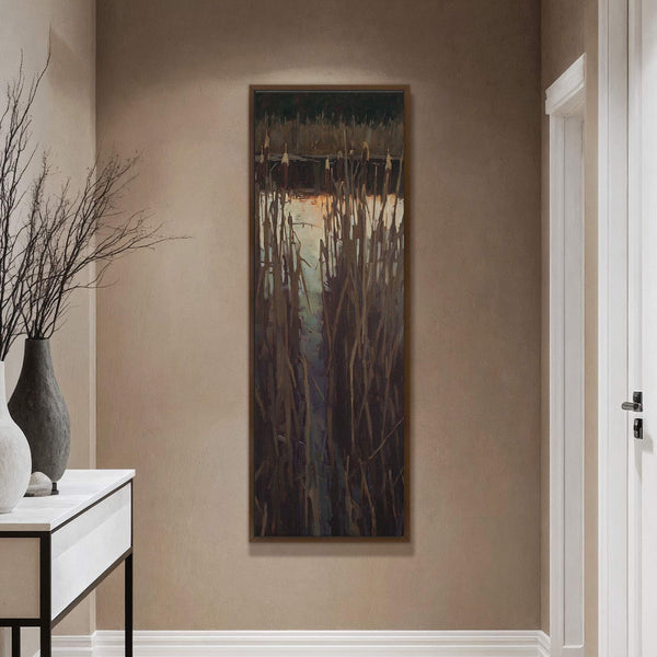 This vertical canvas wall art print displays a painterly depiction of cattails by a body of water, featuring a muted palette and impressionistic style, framed in brown on a taupe.