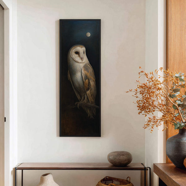 This vertical, dark-toned painting print of a barn owl under a moonlit sky is framed in black and hangs on a white wall above a wooden console table.