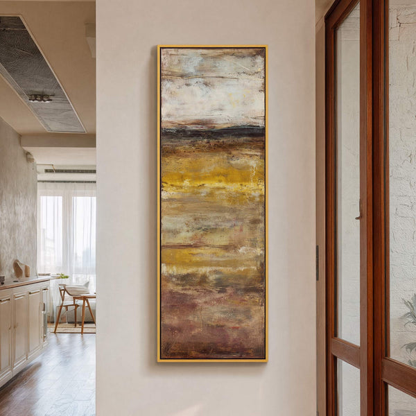 This vertical canvas wall art features an abstract landscape with a palette of warm yellows, browns, and whites, framed in gold and displayed on a beige wall, with a wooden.