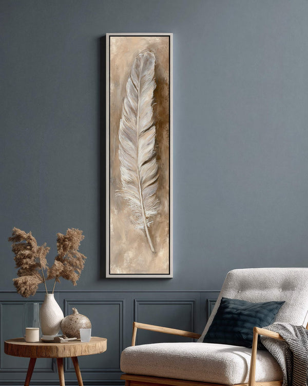 This vertical canvas print features a textured white feather in an earthy palette, displayed in a white floater frame on a dark blue paneled wall beside a plush armchair.
