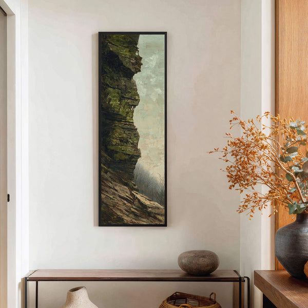 This vertical canvas wall art displays a moody, textured painting of a rocky cliff face with moss and bare trees against a pale sky, framed in black, and hangs above.
