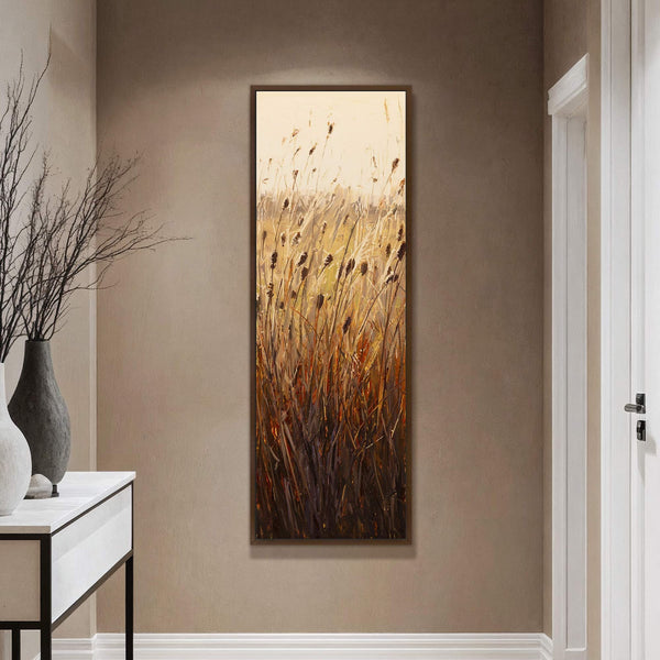 This vertical canvas wall art features a textured, impressionistic painting of tall reeds with a warm, golden palette, framed in dark wood and displayed in a hallway with a textured.