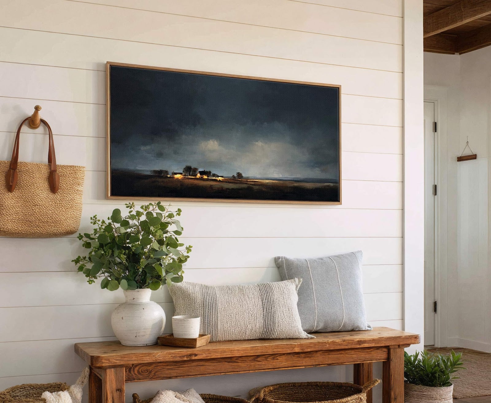 A horizontal canvas wall art depicts a dimly lit, moody landscape with a rustic farmhouse illuminated by warm lights, set against a stormy sky, framed in natural wood and displayed.