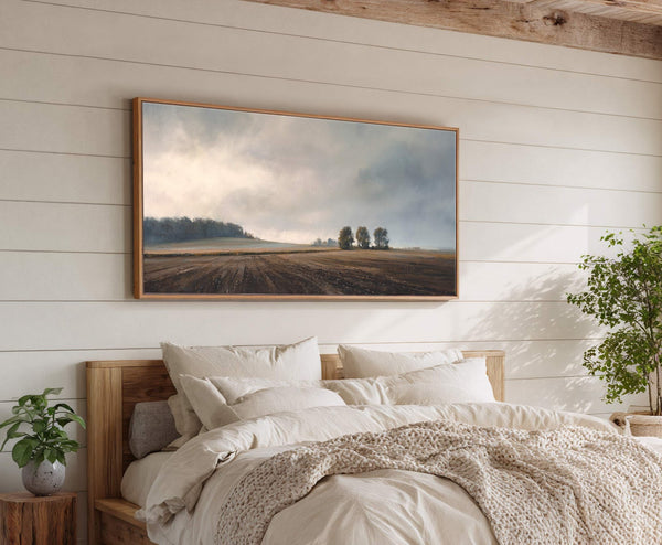 This horizontal canvas print features a muted landscape of a plowed field under a hazy sky with trees in the distance, framed in natural wood, hung above a bed with.