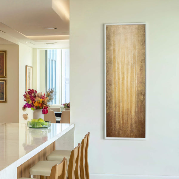 This vertical canvas wall art features an abstract, textured design in warm, golden hues, framed in white and displayed on a pale wall in a modern room with a bar-height.