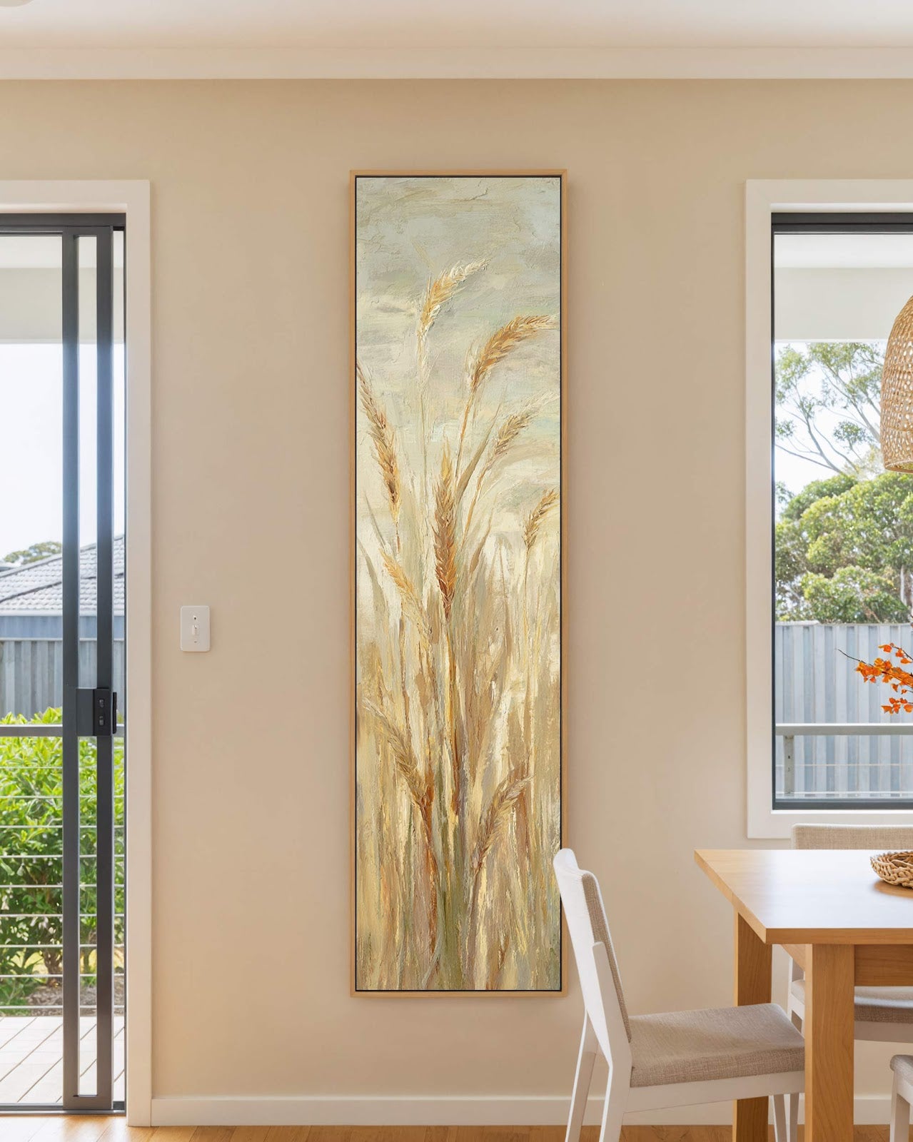 This vertical painting print depicts textured golden wheat in an impressionist style, housed in an oak floater frame on a beige dining room wall beside a wooden table.