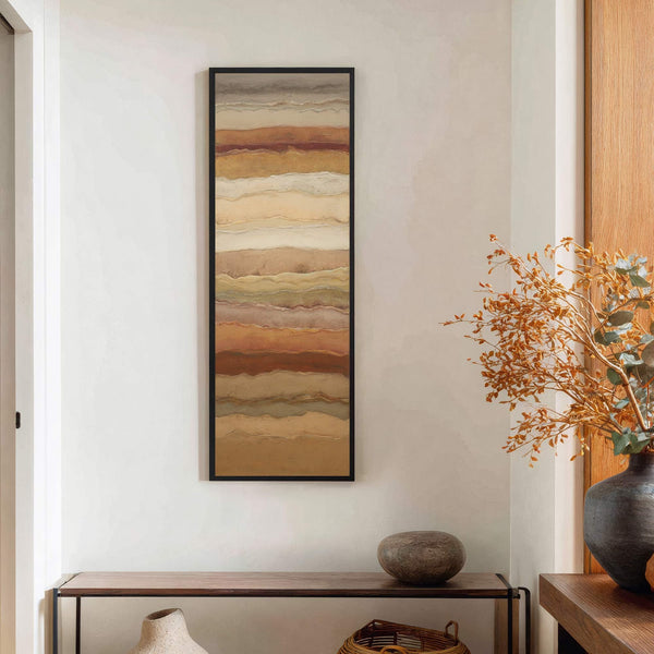 This vertical, framed canvas wall art features an abstract landscape with earthy brown, cream, and rust tones. It hangs on a textured white wall above a wooden console table with.