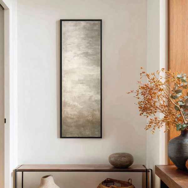 A vertical canvas print with a muted gray and beige abstract palette is framed in black and hangs on a textured white wall above a dark wood console table.
