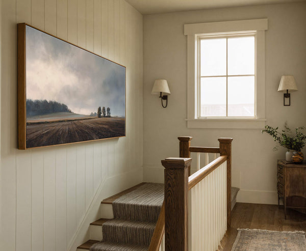 This horizontal canvas wall art features a moody landscape of a tilled field and distant trees under a cloudy sky, framed in natural wood, displayed on a light wood paneled.