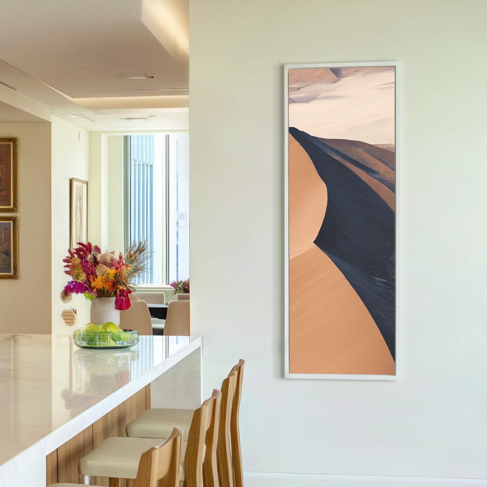 A vertical canvas print of a desert landscape with a dark, curved shadow across tan dunes, framed in white, hangs on a pale wall above a modern kitchen island with.