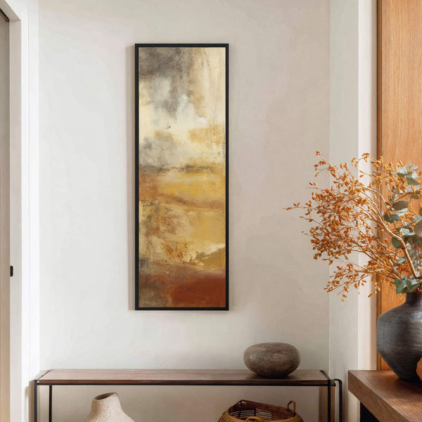 This abstract canvas wall art features a warm earthy palette of ochre, rust, and cream textures, framed in black and displayed vertically on a textured off-white wall above a wooden.