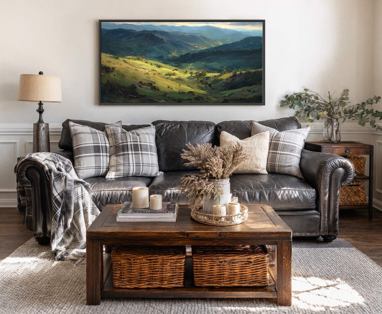A horizontal canvas wall art print with a painterly style depicts rolling green hills bathed in golden sunlight under a hazy blue sky, displayed above a dark leather sofa in.