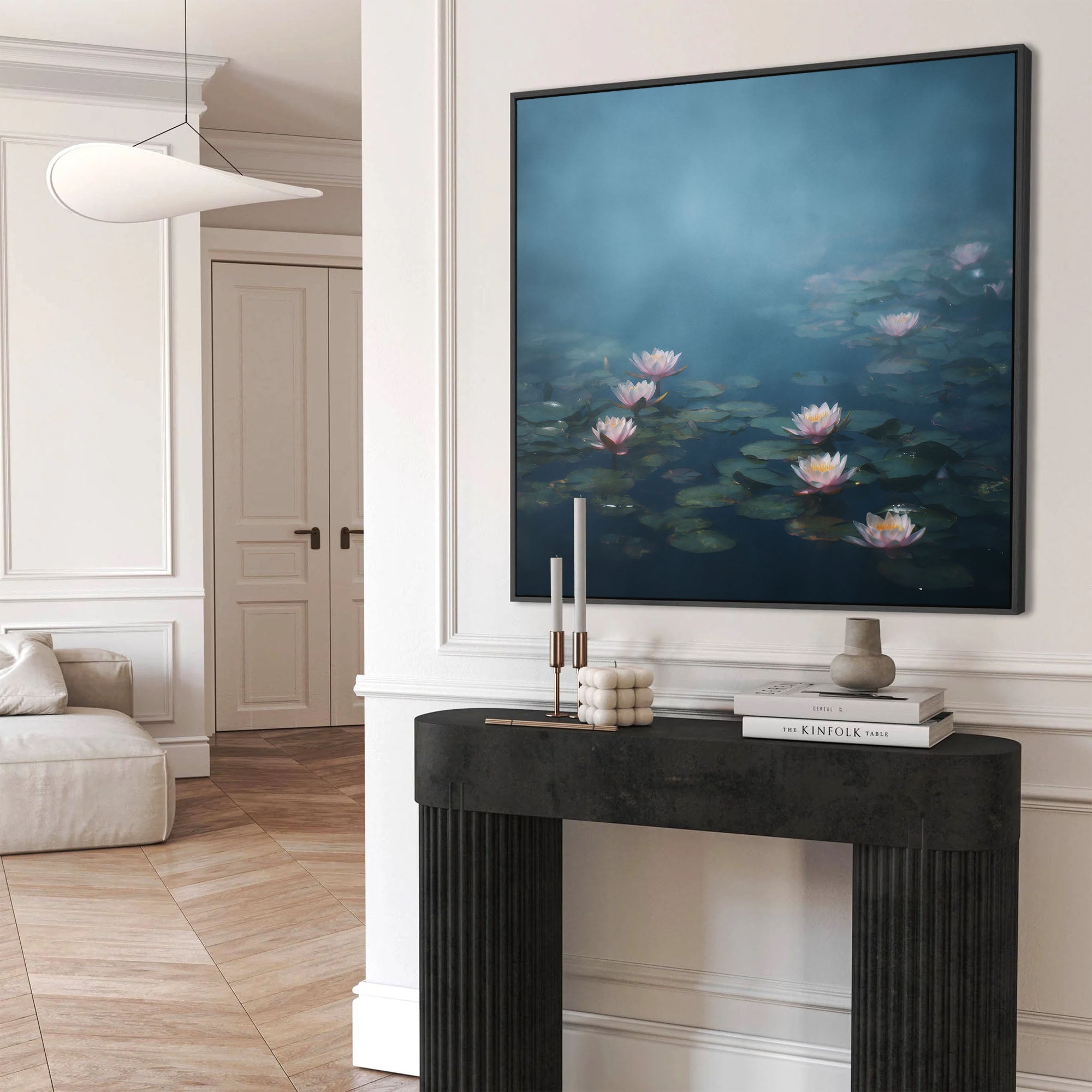 Large framed wall art print depicting serene pink and white water lilies floating on a dark pond under a hazy, ethereal blue mist, hung above a modern black console table in a minimalist white entryway.